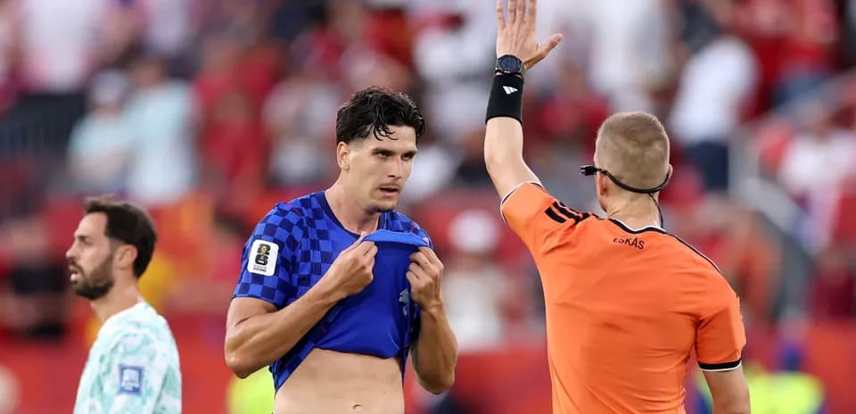 Futbolchi hakamga murojaat qilmoqda, boshqa futbolchi orqada